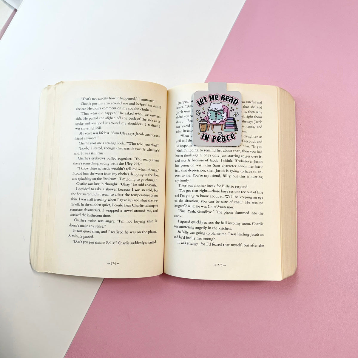 An open vintage book with printed text on the pages, featuring a decorative bookmark or sticker that reads 'LET ME READ IN PEACE' with an illustration of a cat reading. The book is photographed from above on a styled background split diagonally between white and pink.