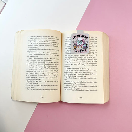 An open vintage book with printed text on the pages, featuring a decorative bookmark or sticker that reads 'LET ME READ IN PEACE' with an illustration of a cat reading. The book is photographed from above on a styled background split diagonally between white and pink.