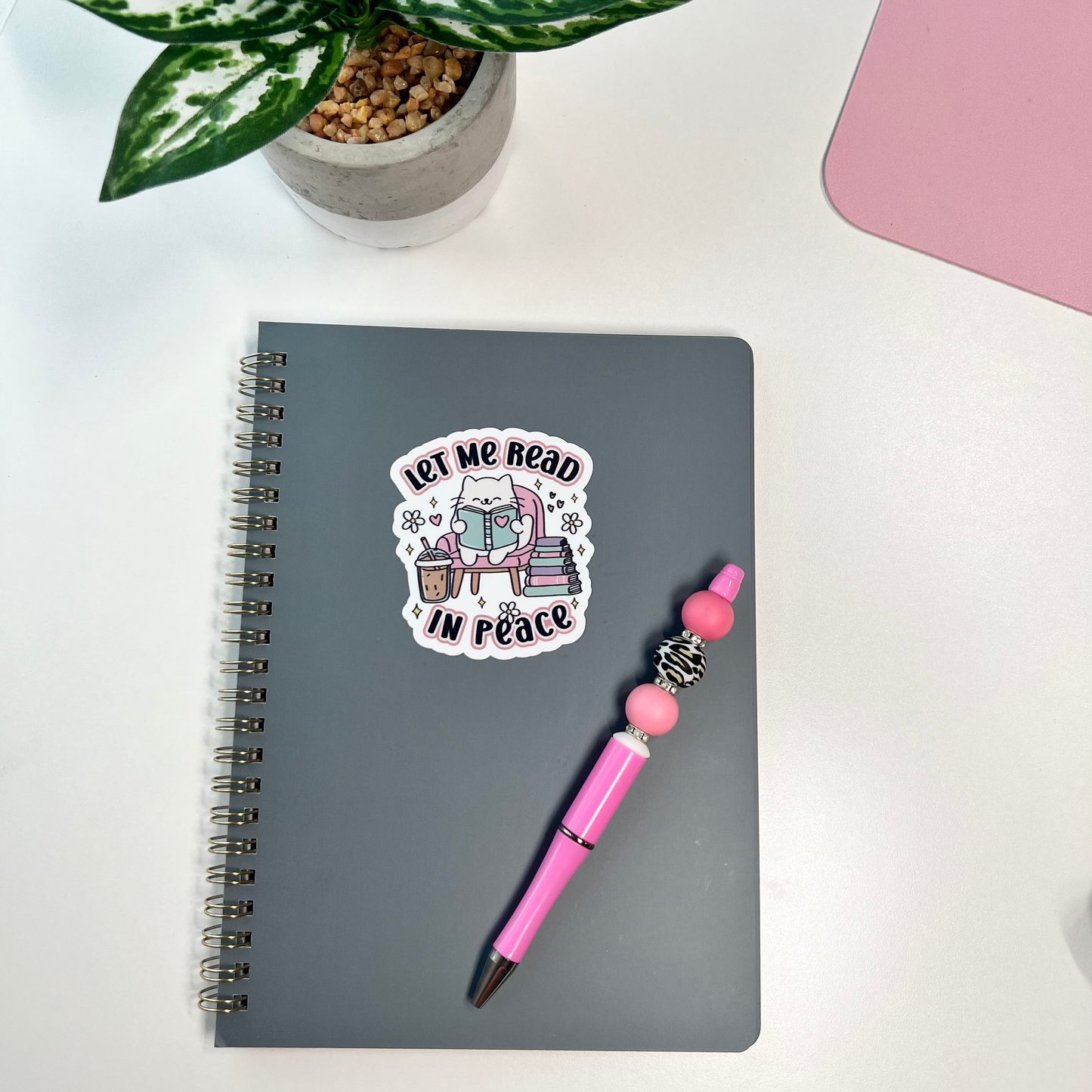 Gray notebook with a sticker, pink pen, and potted plant on a white surface