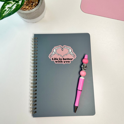 Notebook with a pink pen and decorative sticker on a white surface with a plant and pink item in the background.