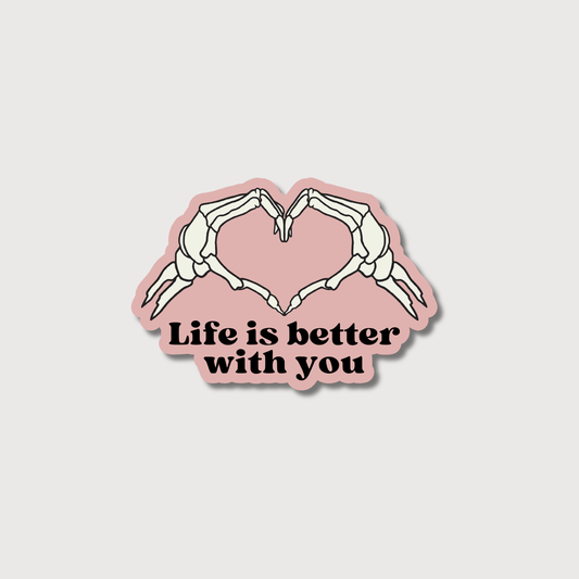 A die-cut sticker featuring two skeletal hands forming a heart shape above the text 'Life is better with you' in black, on a dusty pink background, centered on a light gray backdrop.