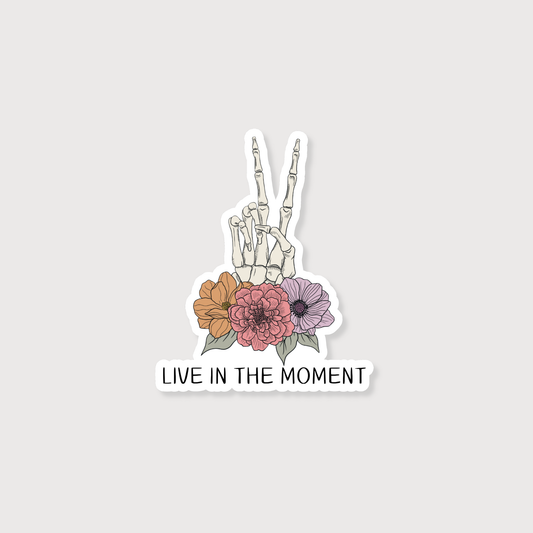 Sticker with skeleton hand making a peace sign and flowers with 'Live in the Moment' text on a light gray background