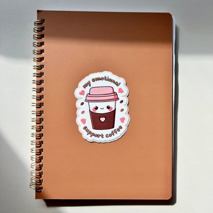 Coffee cup with a heart in the middle with hearts and coffee beans around and the words my emotional support coffee on top of a notebook