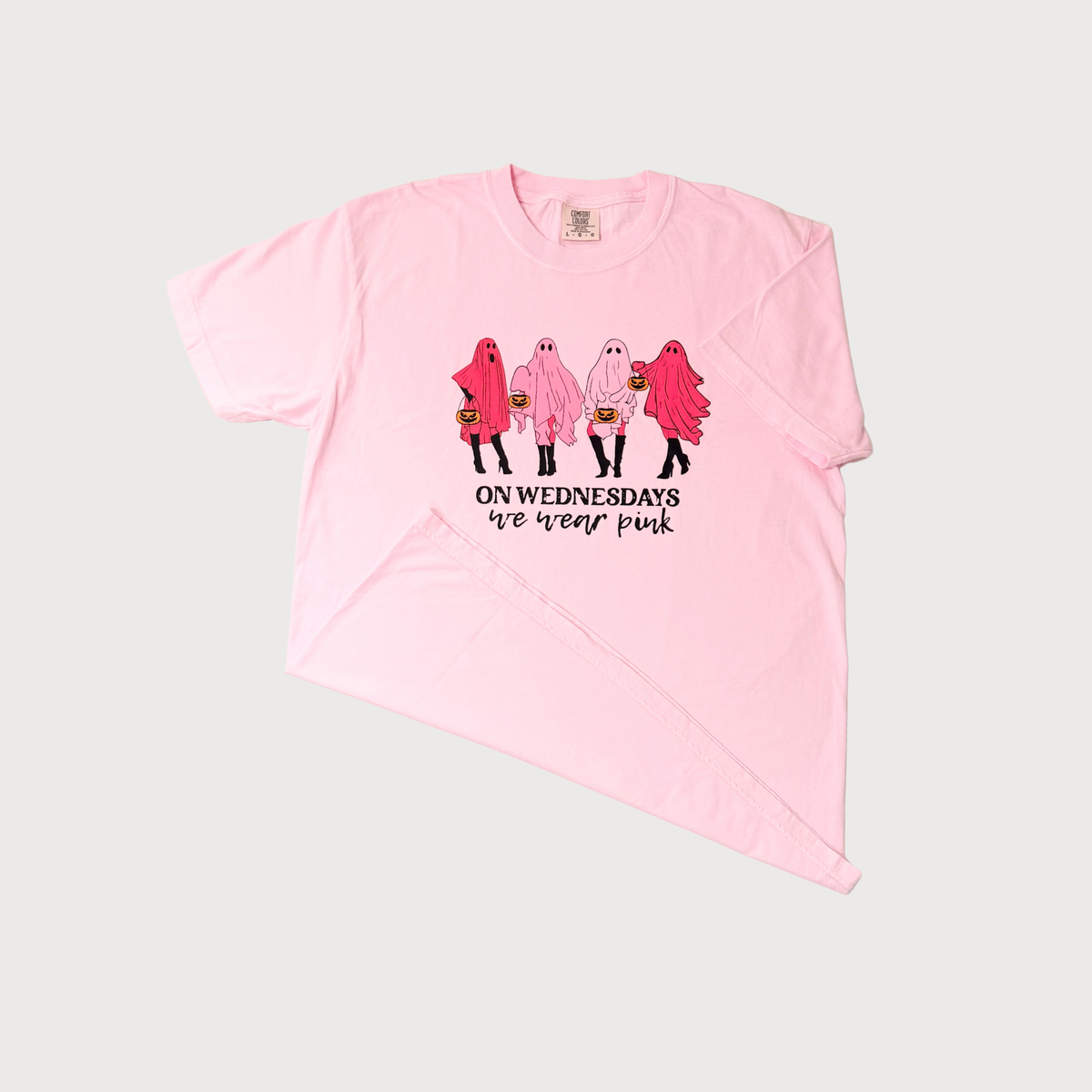 A light pink short-sleeve t-shirt with a graphic of four cartoon ghosts in pink, white, and red costumes holding pumpkins, with the text 'ON WEDNESDAYS we wear pink' printed below. The t-shirt is laid flat on a light gray background.