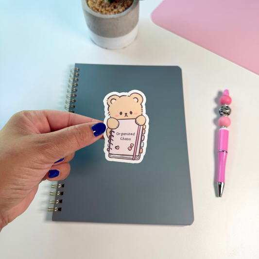 A hand with blue nail polish holds a sticker featuring a cute teddy bear holding a notebook with the words 'Organized Chaos' next to a gray spiral notebook on a white desk. A pink pen, a potted plant, and a pink desk mat are visible in the background.
