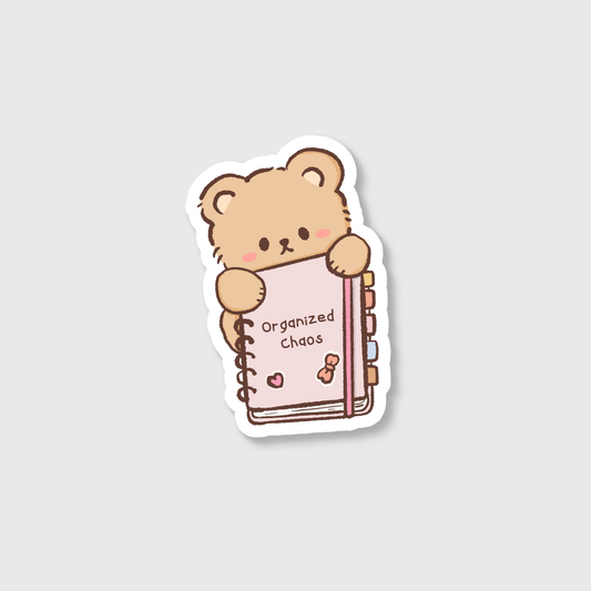 A die-cut sticker featuring a cute cartoon teddy bear holding a pink spiral notebook with the words 'Organized Chaos' on the cover, shown on a light gray background.
