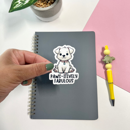 Notebook with a sticker of a dog and text 'PAWS-itively Fabulous' on a desk with a plant and pen.