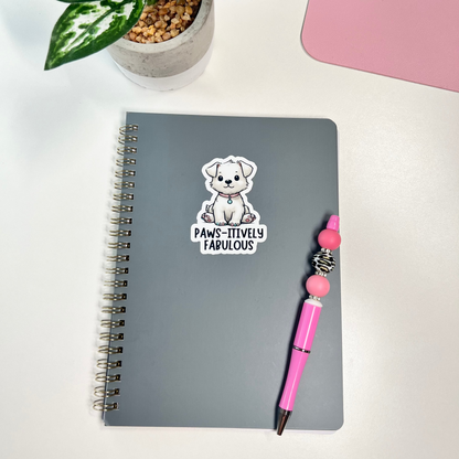 Gray notebook with a sticker of a bear and text, pink pen, and potted plant on a white surface