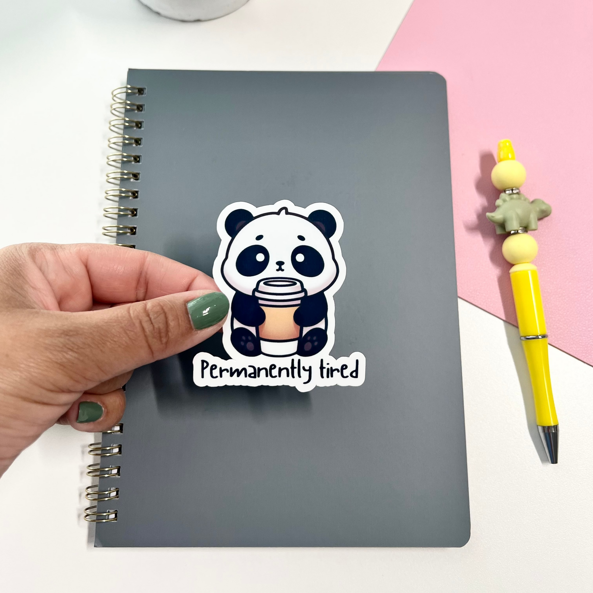 Hand holding a sticker with a panda design on a notebook with a pink and white background.
