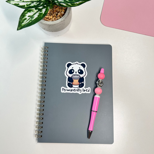 Notebook with a panda sticker and pink pen on a white surface with a plant and pink item in the background