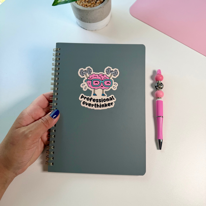 A gray spiral-bound notebook with a cartoon brain sticker that reads 'Professional Overthinker' is held by a hand on a white desk. A pink pen and a small potted plant are nearby.