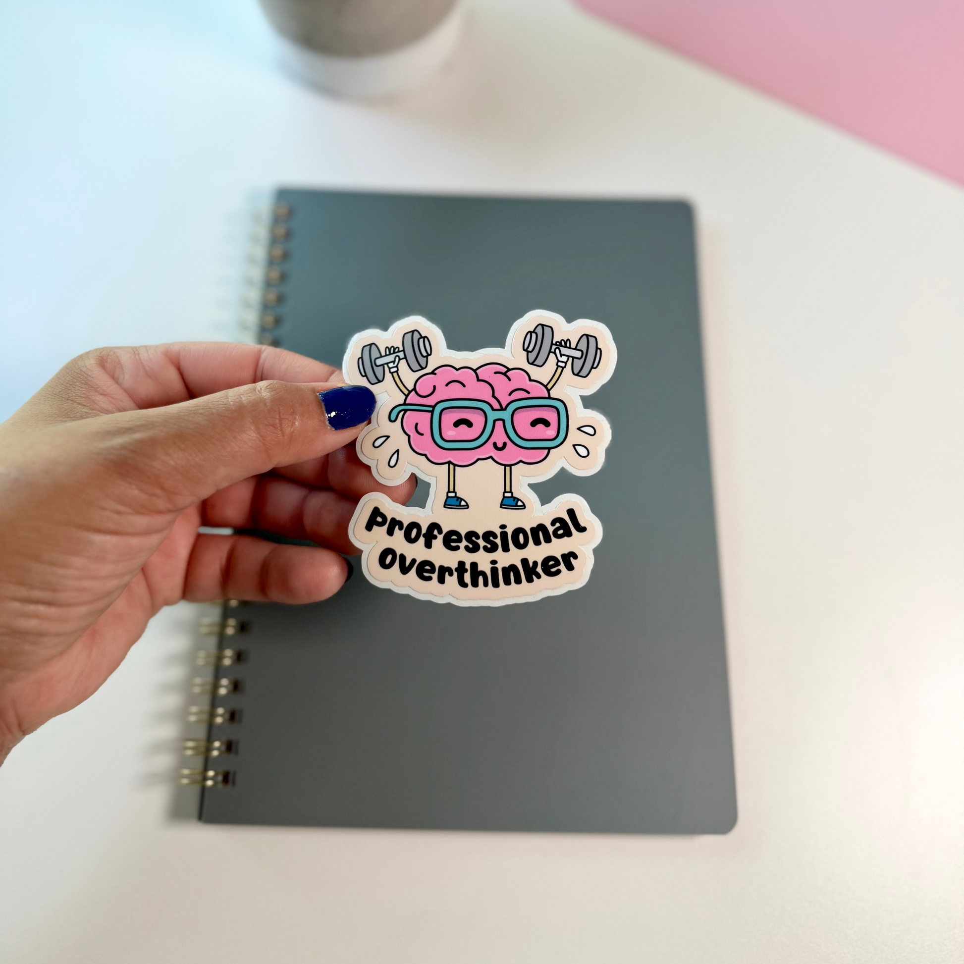 A hand with dark blue nail polish holds a die-cut sticker above a gray spiral notebook on a white desk. The sticker features a cartoon pink brain wearing glasses and lifting barbell weights, with the text 'Professional overthinker'.