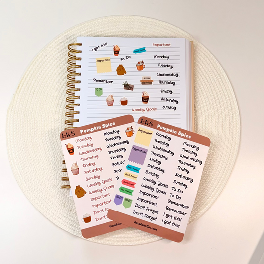 A spiral-bound planner notebook and two sticker sheets with colorful daily habit and mood stickers, arranged on a round woven placemat on a white table.