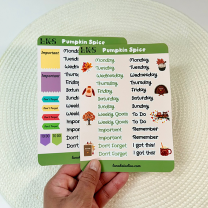 A hand holding a small printed planner insert titled 'Pumpkin Spice' with colorful labels and autumn-themed illustrations, photographed over a white woven placemat on a white tabletop.