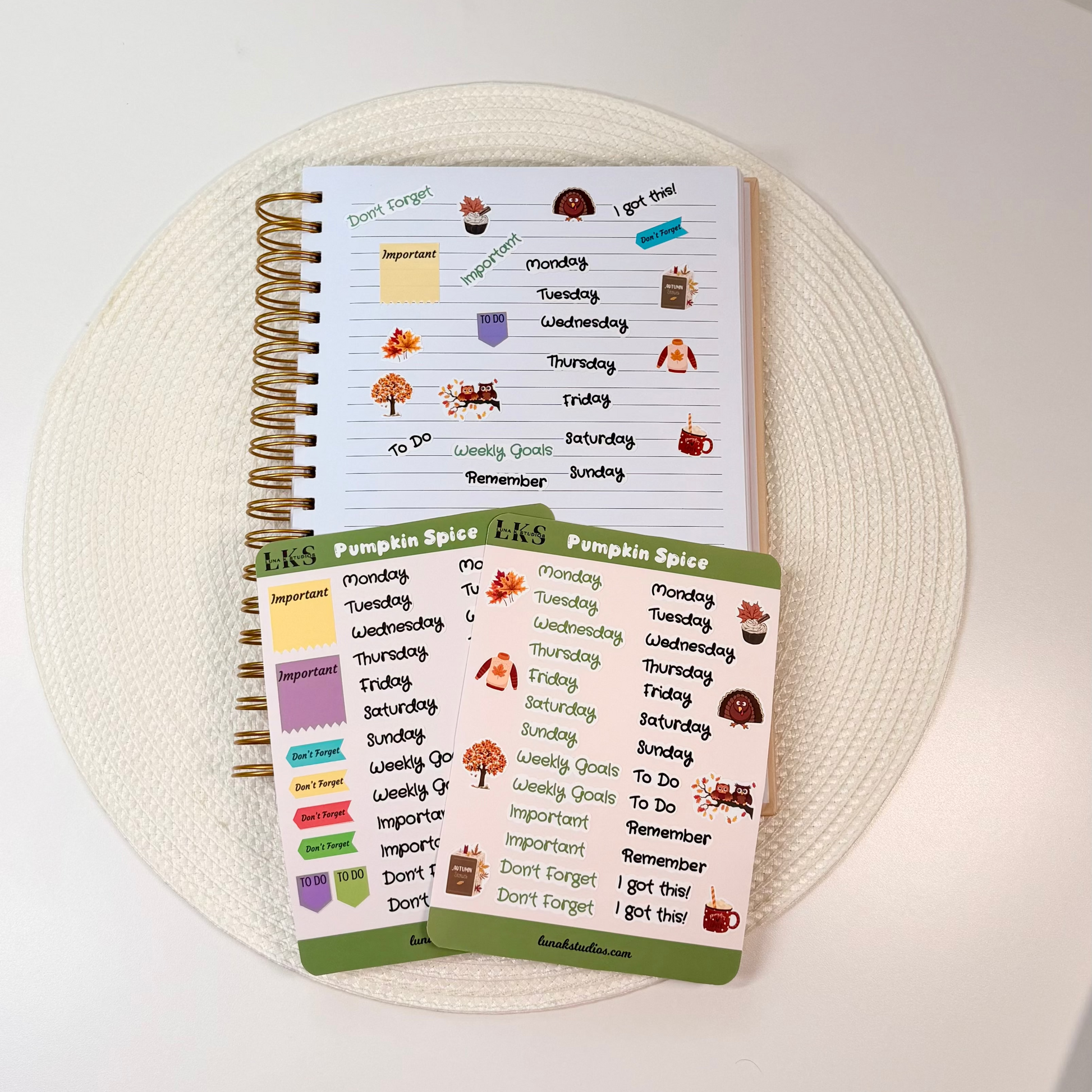 A spiral-bound planner and matching sticker sheets with autumn-themed labels, arranged on a round woven placemat on a white tabletop.