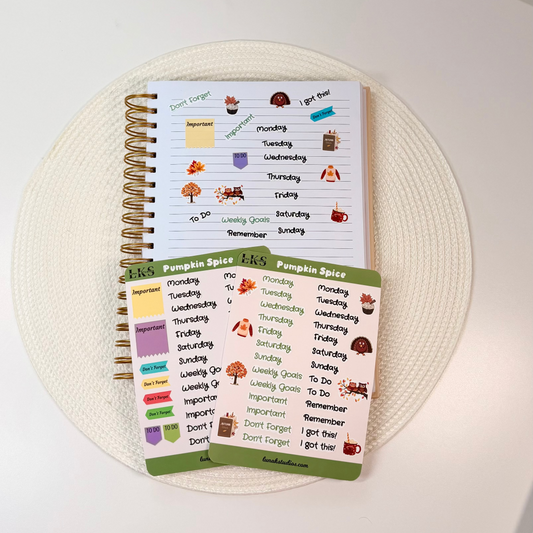A spiral-bound planner and matching sticker sheets with autumn-themed labels, arranged on a round woven placemat on a white tabletop.