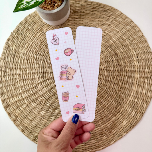 A hand with blue nail polish holds two decorative paper bookmarks or tags with a pink and white grid pattern and cute illustrations, set against a woven round placemat with a potted plant in the background.
