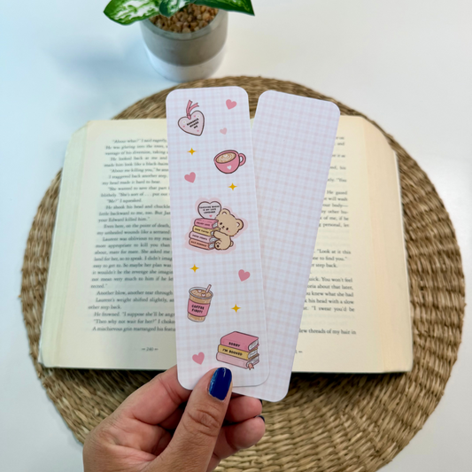 A hand with blue nail polish holds two decorative paper bookmarks featuring cute illustrations of a bear, coffee cups, and hearts on a white grid background. The bookmarks are positioned over an open book on a woven placemat, with a small potted plant in the background.