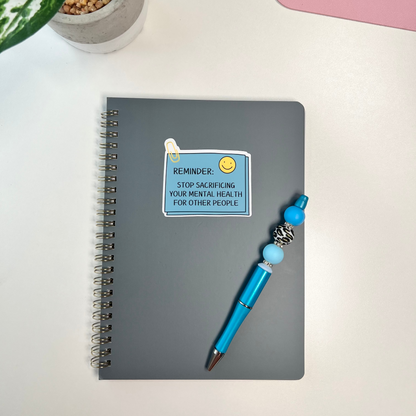 Gray notebook with a blue pen, motivational sticker, and plant on a white surface