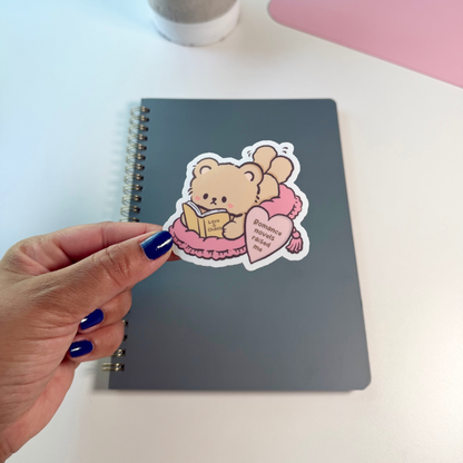A hand with blue nail polish holds a die-cut sticker featuring a cute cartoon teddy bear reading a book and holding a pink heart, placed on top of a dark gray spiral notebook. The scene is set on a white desk with a small potted plant and a pink desk mat in the background.