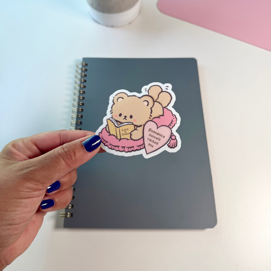 A hand with blue nail polish holds a die-cut sticker featuring a cute cartoon teddy bear reading a book and holding a pink heart, placed on top of a dark gray spiral notebook. The scene is set on a white desk with a small potted plant and a pink desk mat in the background.