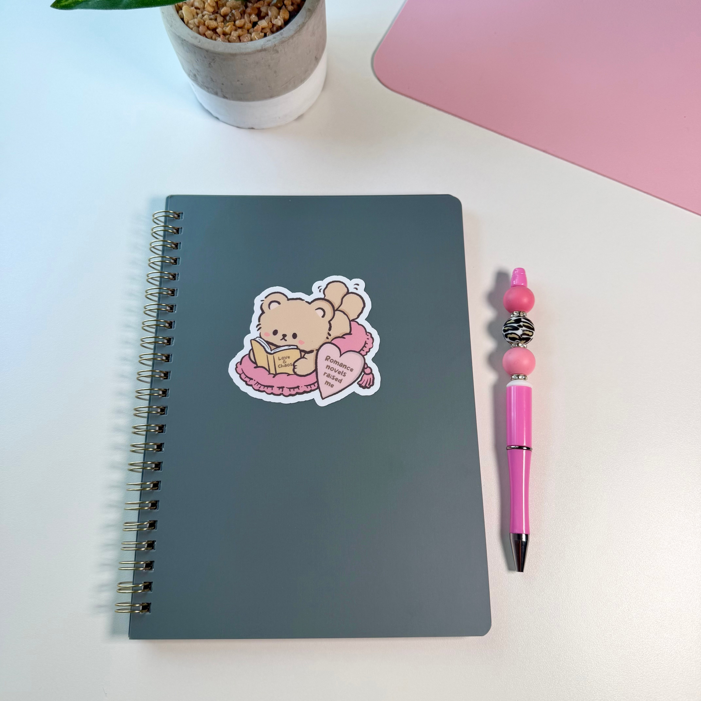 A dark gray spiral-bound notebook with a cute teddy bear sticker, displayed on a white desk next to a pink pen and a potted plant.