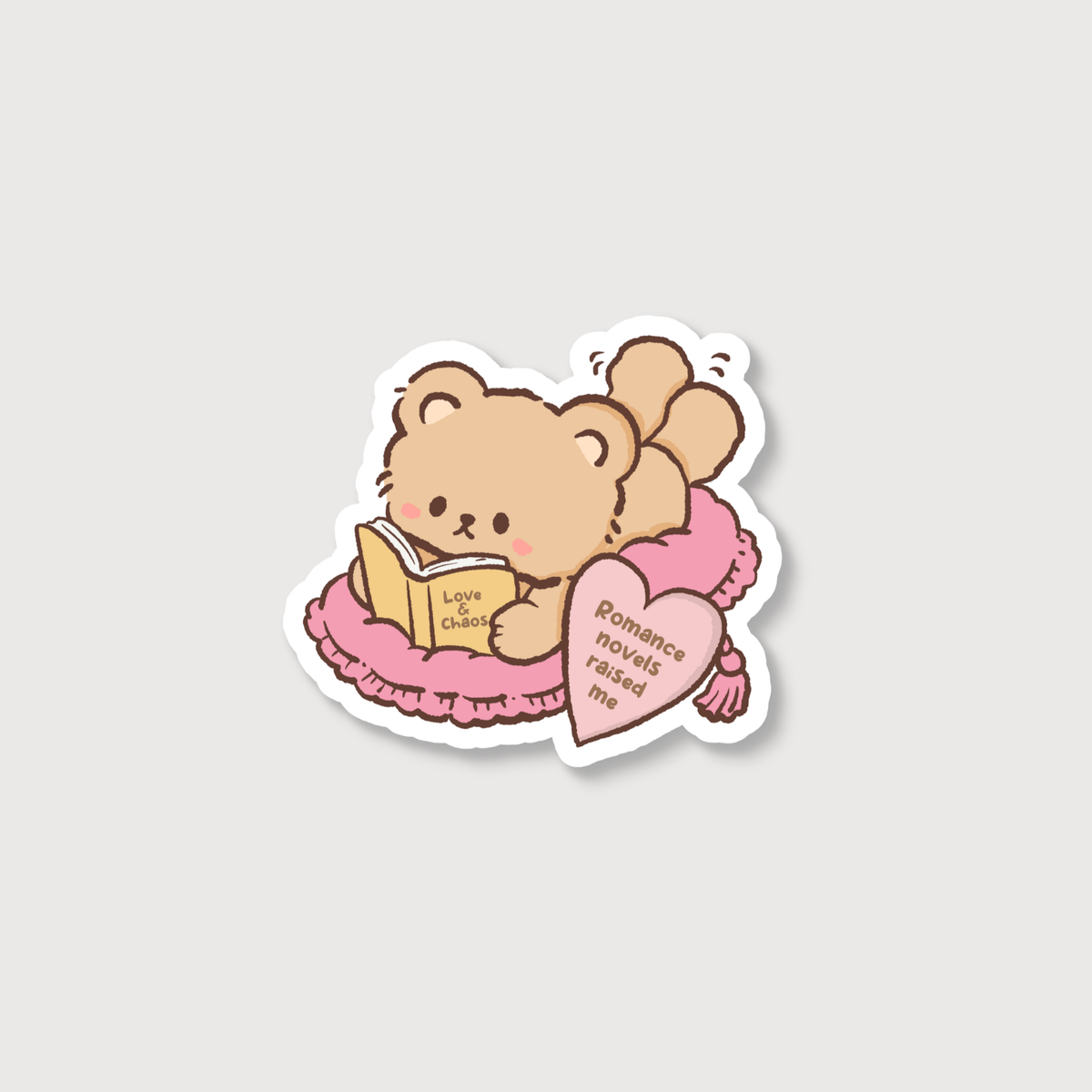 A die-cut sticker featuring a cute cartoon teddy bear reading a book on a pink bed, with a pink heart-shaped pillow that reads 'Romance novels raised me'.
