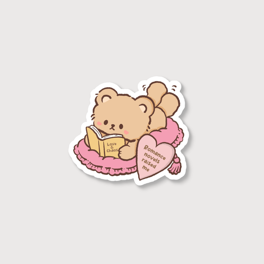 A die-cut sticker featuring a cute cartoon teddy bear reading a book on a pink bed, with a pink heart-shaped pillow that reads 'Romance novels raised me'.