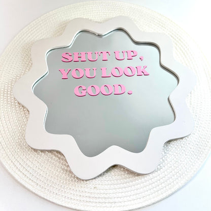 A scalloped white mirror with pink text reading 'SHUT UP, YOU LOOK GOOD.' placed on a round woven white placemat against a white background.