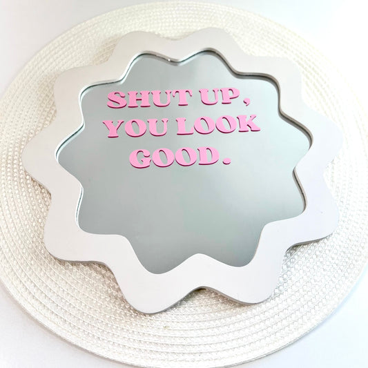 A scalloped white mirror with pink text reading 'SHUT UP, YOU LOOK GOOD.' placed on a round woven white placemat against a white background.