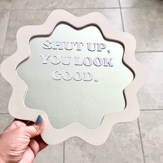 A hand with blue nail polish holds a scalloped white mirror with the phrase 'SHUT UP, YOU LOOK GOOD.' embossed in the reflective surface, set against a gray tiled floor background.