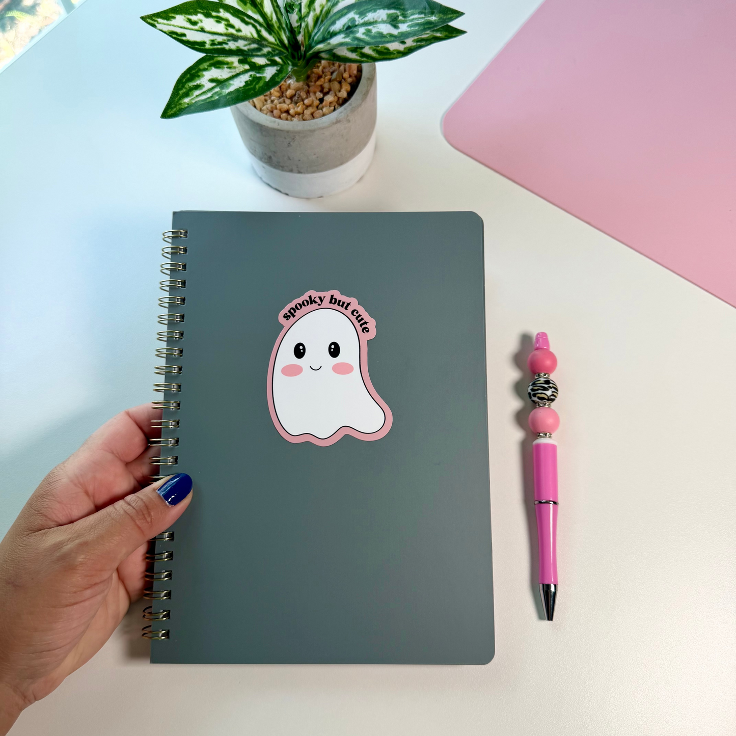 A dark gray spiral notebook with a cute ghost sticker that reads 'Spooky but cute' is held by a hand with blue nail polish. The notebook is styled on a white desk next to a pink pen and a small potted plant.