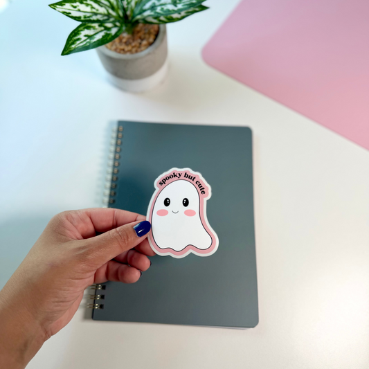A hand with blue nail polish holds a cute ghost sticker that says 'spooky but cute' above a gray spiral notebook on a white desk, with a potted plant and a pink laptop in the background.