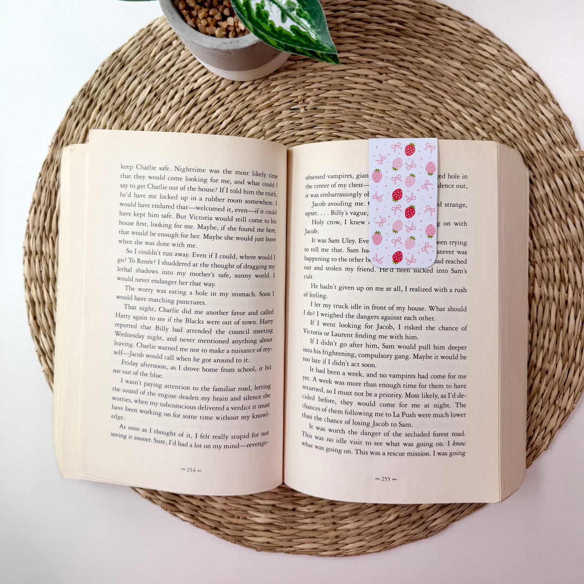 An open book spread displayed on a round woven mat, with a small potted green plant behind it. The right page has a pink bookmark featuring a red polka-dot pattern.