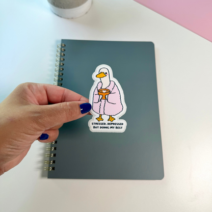 A hand with dark blue nail polish holds a die-cut sticker featuring a cartoon duck in a pink robe with the text 'STRESSED, DEPRESSED BUT DOING MY BEST'. The sticker is placed on a dark gray spiral notebook on a white desk, with a potted plant and a pink laptop visible in the background.