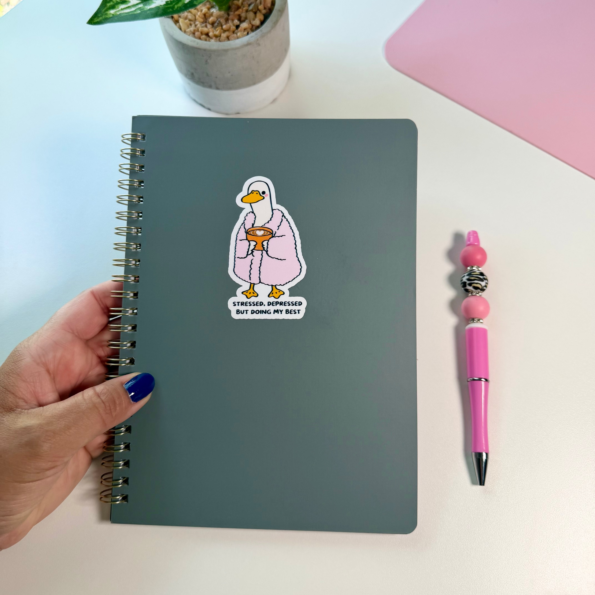 A hand with blue nail polish holds a dark gray spiral notebook with a cartoon duck sticker on the cover. The notebook is on a white desk next to a pink pen and a potted plant.