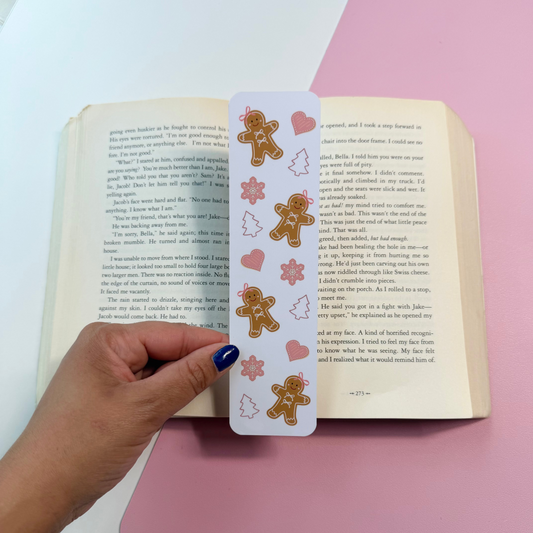 A hand with dark blue nail polish holds a white bookmark featuring gingerbread men and hearts over an open book, set against a split pink and white background.