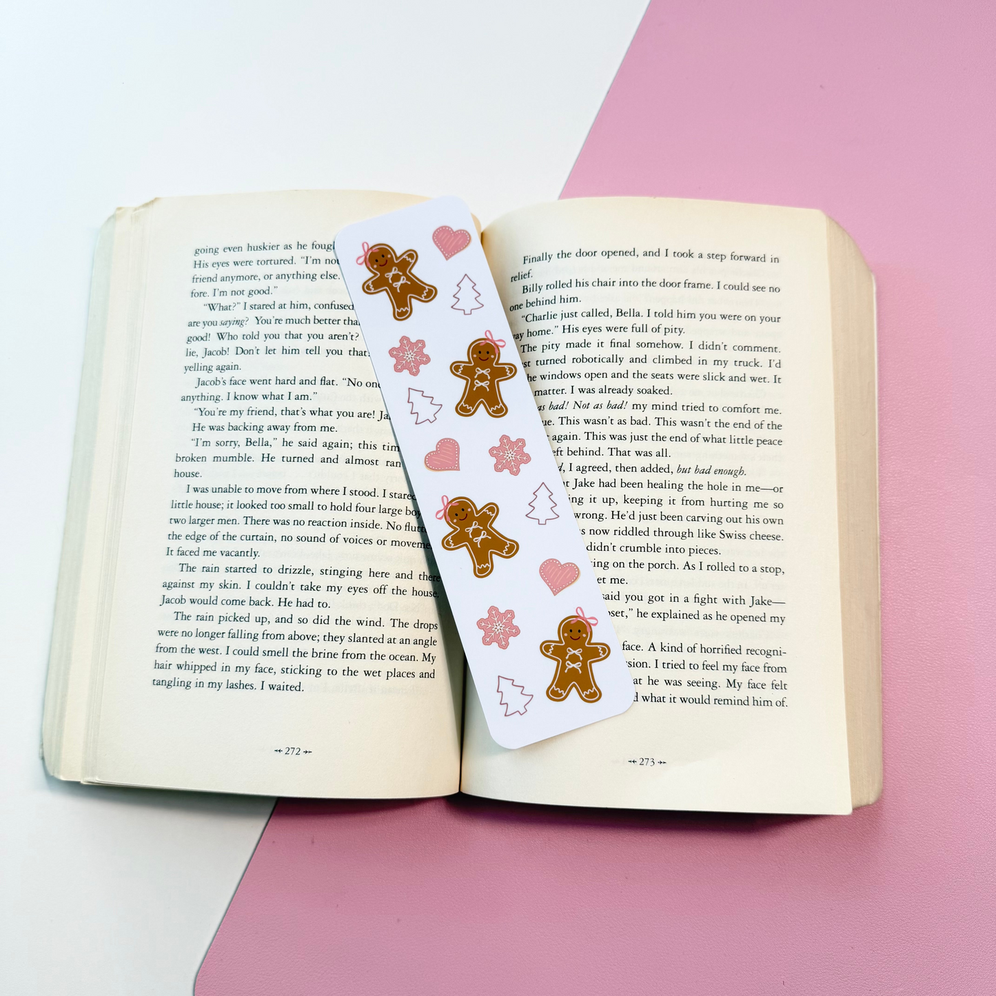 A white paper bookmark with gingerbread men and pink hearts printed on it, laid across an open book on a pink and white surface.
