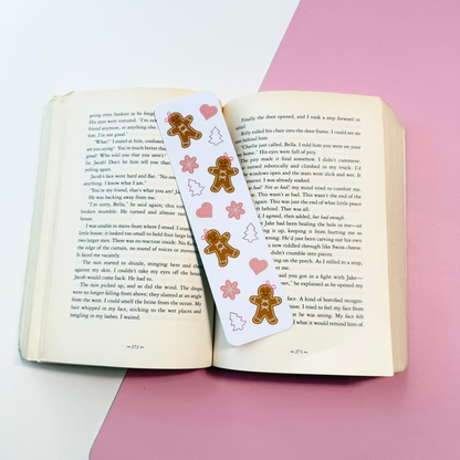 A white paper bookmark with gingerbread men and pink hearts printed on it, laid across an open book on a pink and white surface.