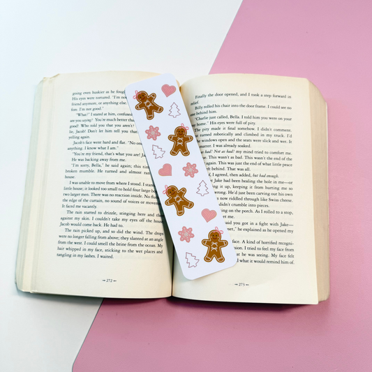 A white paper bookmark with gingerbread men and pink hearts printed on it, laid across an open book on a pink and white surface.