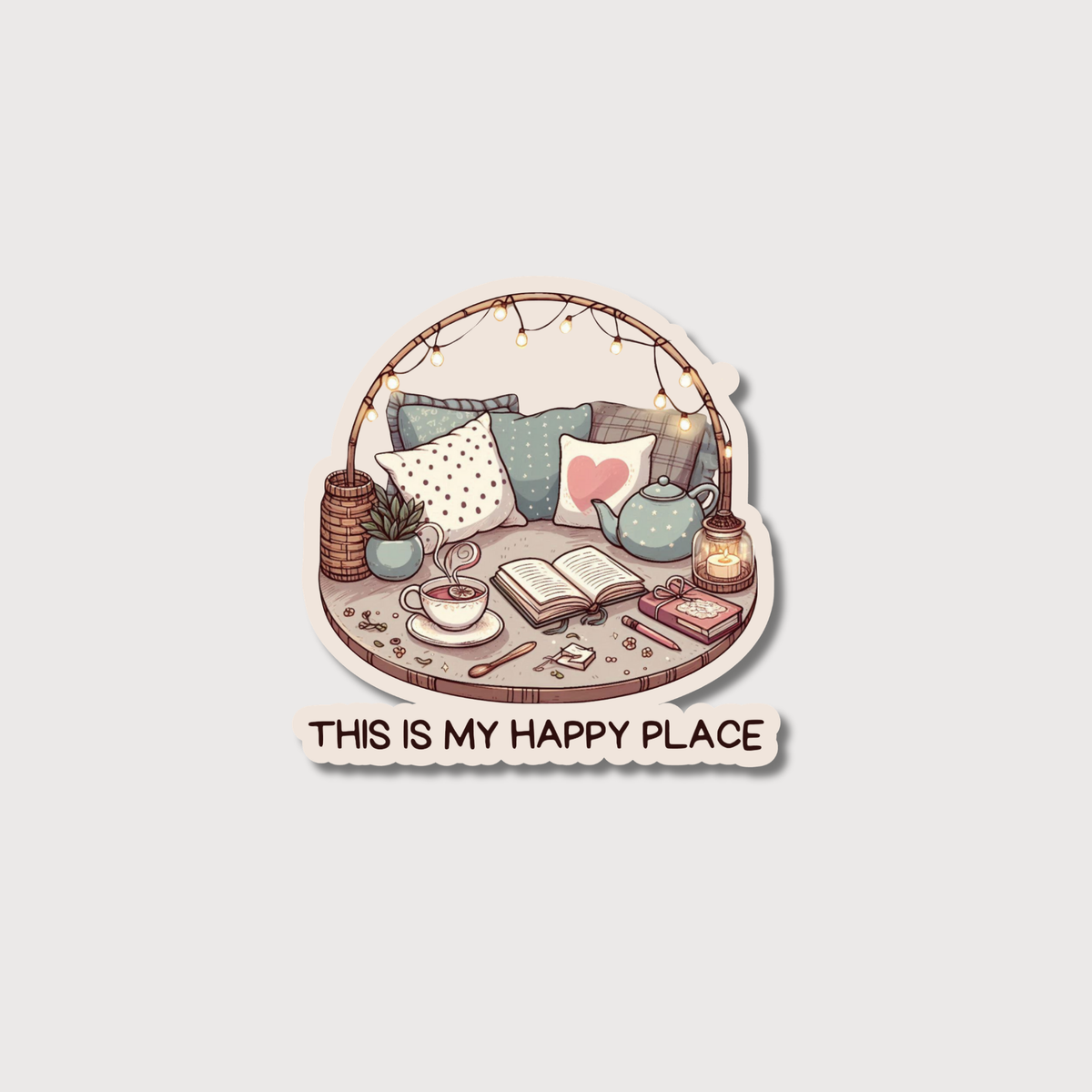 A die-cut sticker featuring a cozy illustrated scene with pillows, a teapot, an open book, a cup of tea, and string lights, with the text 'THIS IS MY HAPPY PLACE' below on a light gray background.