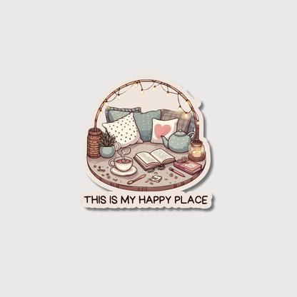 A die-cut sticker featuring a cozy illustrated scene with pillows, a teapot, an open book, a cup of tea, and string lights, with the text 'THIS IS MY HAPPY PLACE' below on a light gray background.