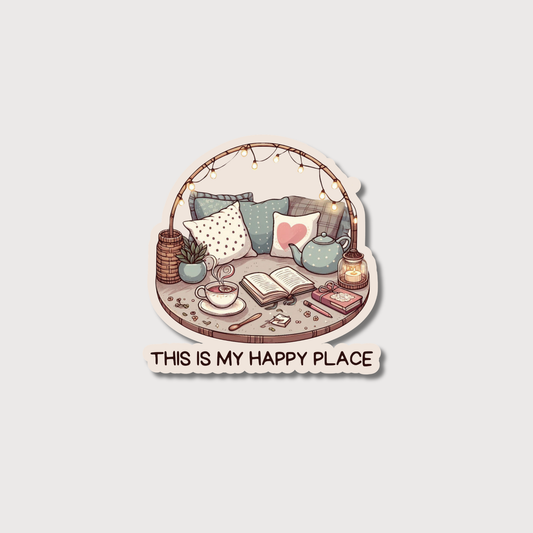 A die-cut sticker featuring a cozy illustrated scene with pillows, a teapot, an open book, a cup of tea, and string lights, with the text 'THIS IS MY HAPPY PLACE' below on a light gray background.