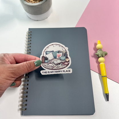 Notebook with a sticker labeled 'This is my happy place' on a white surface with a plant and pen.