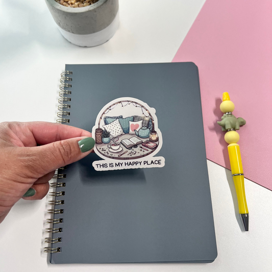 Notebook with a sticker labeled 'This is my happy place' on a white surface with a plant and pen.