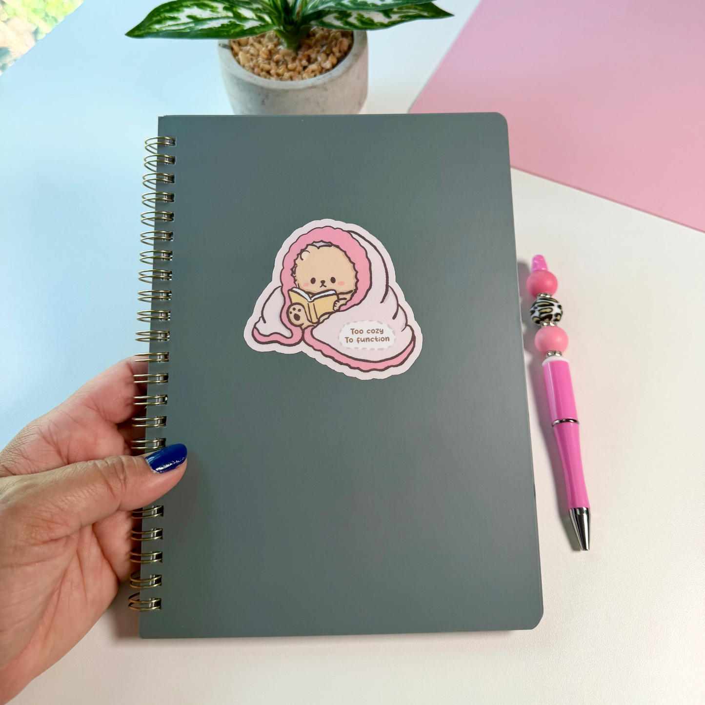 A dark gray spiral-bound notebook with a cute pink sticker of a bear wrapped in a blanket. A pink pen and a small potted plant sit on a white desk next to a pink background.