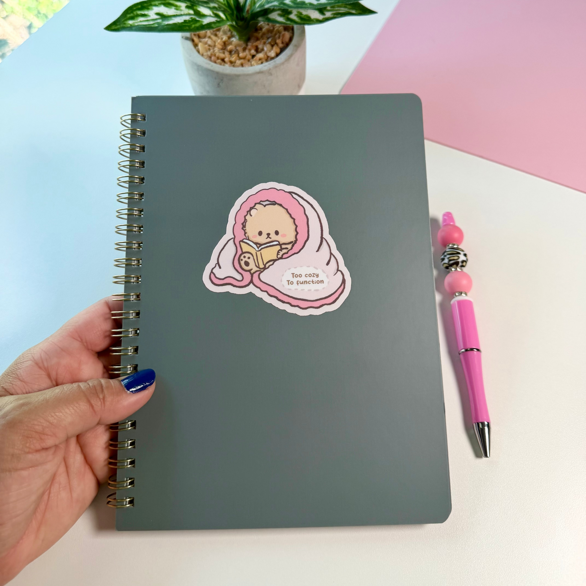 A dark gray spiral-bound notebook with a cute pink sticker of a bear wrapped in a blanket. A pink pen and a small potted plant sit on a white desk next to a pink background.