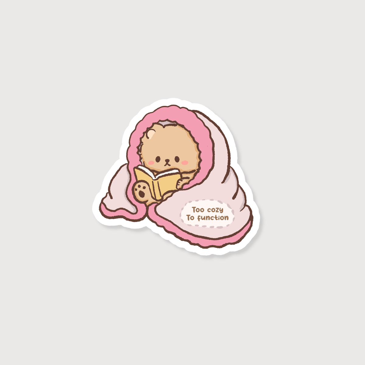 A die-cut sticker featuring a cute cartoon illustration of a small beige character wrapped in a pink blanket and reading a book, with the text 'Too cozy to function' on a light gray background.