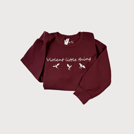A maroon long-sleeve crewneck sweatshirt with 'Violent Little Thing' and three small white dragon illustrations printed on the chest, displayed flat against a light gray background.