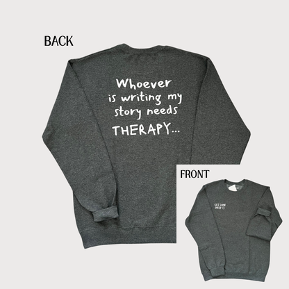 Dark heather gray crewneck sweatshirt shown from both front and back on a light gray background. The back view has large white handwritten-style text reading 'Whoever is writing my story needs THERAPY.